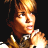 Pixelated close-up of a person with light brown hair and fair skin wearing a dark outfit with white accents, set against a black background.