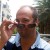 Pixelated static image of a person holding a glass near their face, wearing dark sunglasses and light-colored clothing against an outdoor background.