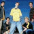 Group of young men in casual clothing posed against blue and white backgrounds, some standing, some seated.