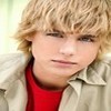 A close-up of a person with tousled blonde hair wearing a red shirt under a light-colored jacket against a blurred background.