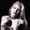 Black-and-white image of a person with long blonde hair wearing a dark choker necklace and holding hands near the chest, displayed in a