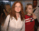 A woman with long red hair in a white jacket and red-and-white striped shirt waves her right hand while standing among other people.