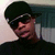 Pixelated image of a person wearing dark cap and shirt, light collar visible, against blurred background with pinkish and blue tones.