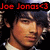 Pixelated image of a person with dark hair and light skin, wearing a white shirt under a dark jacket, with red text reading "Joe Jonas" above the head.