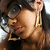 A person with long dark hair wears large round glasses and gold earrings, smiling slightly while looking downward.