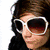 A static image of a person with dark hair wearing white-framed sunglasses and a red jacket over black clothing, seated in what appears to be a vehicle interior.