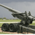 A large military artillery piece with a long barrel is positioned on a grassy field under a cloudy sky.