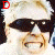 A pixelated image of a person with light hair wearing dark sunglasses and a black top, holding a brown object near their face.