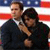 Two people in dark suits stand side-by-side against a red, white, and blue striped background with white stars on blue.