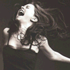 Black-and-white image of a woman with long hair in a dark dress, head tilted back, mouth open in a scream or laugh, arms extended against a dark background.