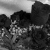 Black-and-white pixelated image of a figure with dark hair and light clothing standing among rocky terrain under a gradient sky.