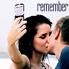 A woman with dark hair holds a flip phone to take a selfie while a man kisses her cheek; the word 'remember' appears above them.