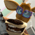 Brown cartoon animal with large ears and blue sunglasses holding a stack of dark brown items against a light background.
