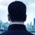 A pixelated image shows the back of a person's head with short dark hair, wearing a dark shirt, against a light blue sky and cityscape background.