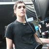 A person in a black t-shirt with a small dark logo on the chest, standing in front of a dark background with a partially visible person holding a camera.