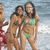 Two women in bikinis stand on a beach with ocean waves behind them; one woman wears a green bikini top and bottom, the other a dark patterned bikini.