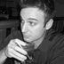 Black-and-white static image of a person with short hair wearing a dark shirt, holding an object near their face in a close-up portrait style.