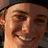 A static image shows a person with light skin and brown hair wearing a dark cap, smiling widely against a black background with white text.