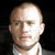 Static pixelated portrait of a bald man with light skin wearing a white shirt against dark background, rendered in low-resolution 256x256 pixels with visible dithering and compression artifacts.