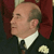 Pixelated profile view of a bald man in a dark suit with a white shirt and dark tie, wearing a light-colored flower on his lapel against a dark red background.