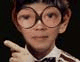 A child wearing large round glasses and a striped shirt points with the right hand against a dark background.
