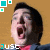 Pixelated static image of a person with dark hair and open mouth against brown background, featuring green square icons in top-left corner and partial text at bottom left.
