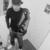 Pixelated grayscale image of a person in dark clothing holding a long object, standing against a light background with partial shapes on the left and right edges.