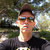 Pixelated static image of a person with short dark hair wearing sunglasses and a dark shirt against a blurred outdoor background.