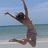 Pixelated static image of a person jumping on a beach with arms raised, wearing dark sunglasses and a striped swimsuit against a blue sky and ocean background.
