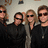 Four people in dark clothing pose together with sunglasses against a light background, accompanied by green pixelated text on black and gray
