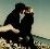 Pixelated static image of two figures in dark clothing embracing on a beach with water and sky background.