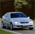 Pixelated image of a light blue sedan with dark blue windows parked on a paved surface, background features dark brown and green foliage.