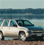 Pixelated image of a white convertible car with dark roof and blue accents parked on a beach near water.
