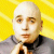 Pixelated static image of a bald person with light skin wearing a gray jacket over a black shirt and brown tie against bright yellow background.