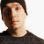 Pixelated close-up of a person's face and upper torso against a white background, wearing dark clothing with visible hair and facial features.
