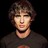 Pixelated static image of a person with short brown hair wearing dark clothing against black background.