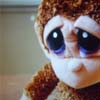 A close-up of a doll with large blue eyes, brown curly hair, and a light-colored face, wearing a brown textured garment against a blurred background.