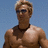 A shirtless man with blonde hair and sunglasses stands against a blue sky background, wearing a black necklace.