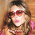 Pixelated image of a person with long brown hair wearing red heart-shaped sunglasses and a pink earring, set against a warm-toned background.