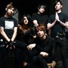 Five people in black clothing pose together against a dark background, with one person wearing a light-colored cap and another with a white square graphic on their shirt.