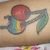 A tattooed arm displays a colorful tattoo of a red fruit with a stem and leaf, a rainbow-colored guitar, and a black musical note.