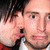 A static image of two people in close proximity, one with dark hair and a red garment, the other with light skin and a white garment, both facing forward.