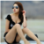 Woman in black bikini with sunglasses, seated on beach, hand near face, long brown hair, light skin tone, blurred background.