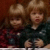 Two pixelated figures with light brown hair, one in dark clothing and the other in red patterned fabric, against a blurred background.