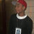 A person wearing a red cap and black sweater with white collar stands against a tiled wall, holding a rectangular object.