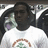 A pixelated static image shows a person with dark skin wearing a white shirt with a brown and green circular design, set against a light background with dark shapes.