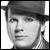 Black-and-white pixelated portrait of a person with short dark hair, wearing a dark cap and light-colored shirt, facing slightly left with neutral expression.