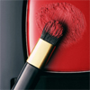 A close-up of a makeup brush with a black handle and gold band resting against a red surface with a circular pattern.