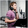 A person in a purple shirt with dark gray sleeves stands in a studio, wearing large black headphones, holding a microphone near their mouth, with a keyboard and microphone stand visible in the background.