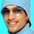 Pixelated close-up of a smiling person with dark hair tied back, light skin tone, and visible teeth against a bright blue background.